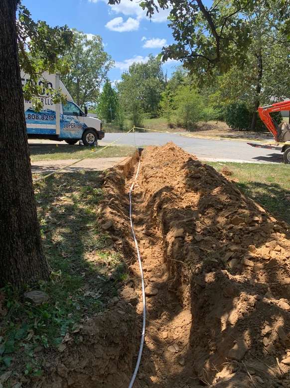 A recently dug ditch in the shade of a tree. A new water line has been laid in the ditch. The Superior Plumbing & Rooter LLC truck is parker in front of the ditch.