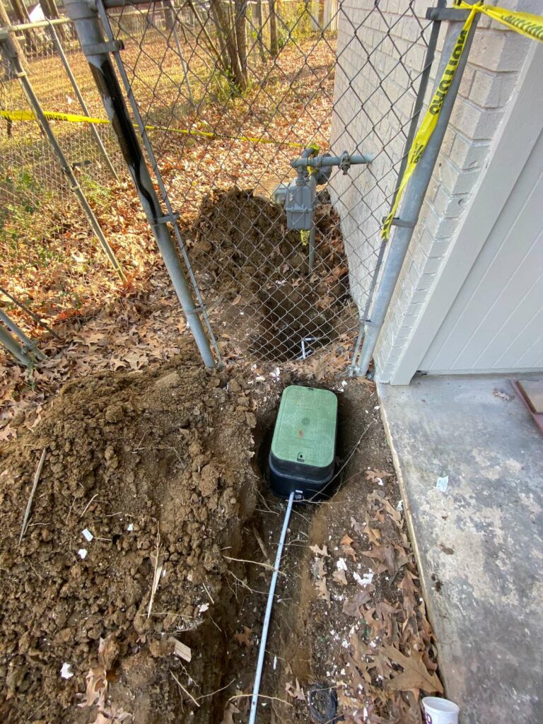 A shallow ditch for an irrigation box with a pipe running out of the box. The box is by a chain link fence that has caution tape around it.