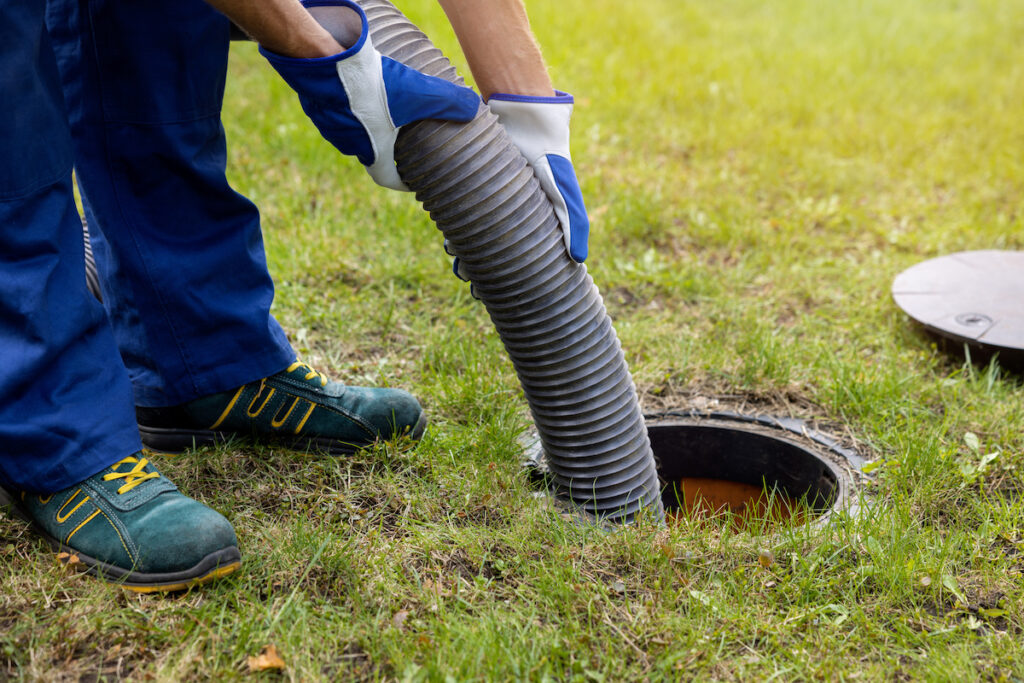 A shot of a man's shoes and blue gloved hands as he pulls a septic tank plastic pipe out of a drain in a green yard.