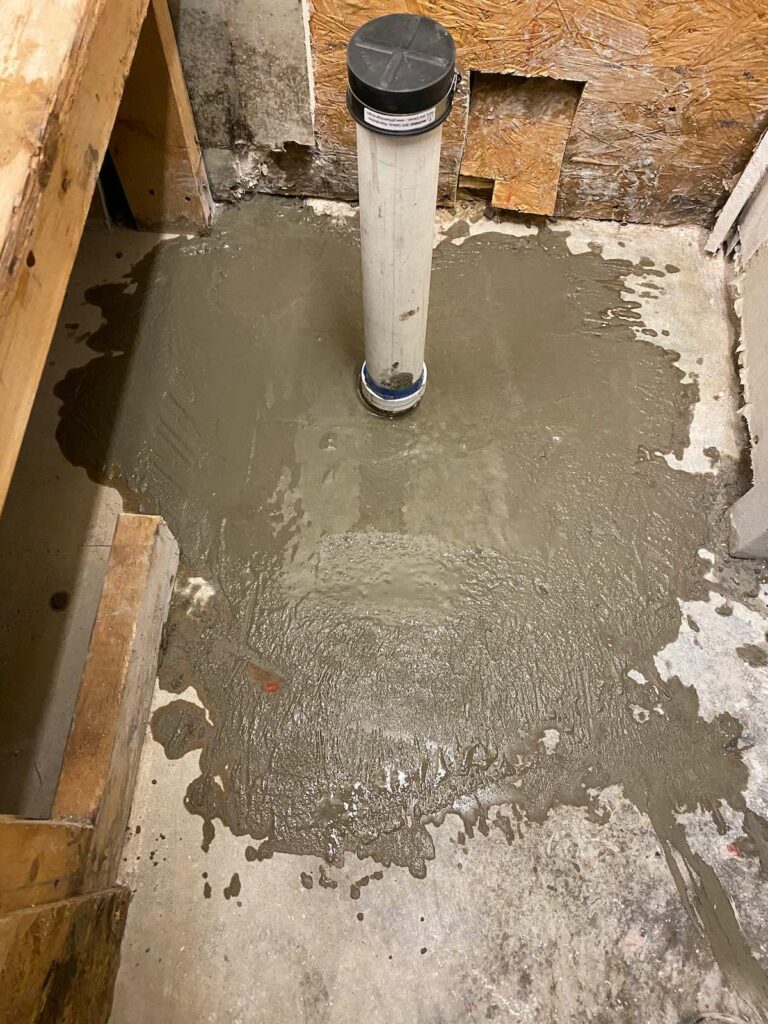 A look down onto a white pipe sticking out fron a concrete floor. Spreading from the bottom of the pipe is muddy water.