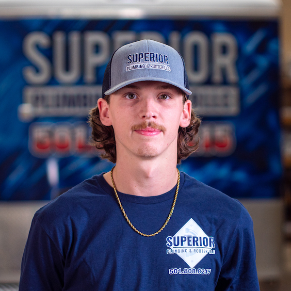 A Superior Plumbing & Rooter LLC intern named Alex. He has a brown mustache and long curly hair sticking out of his ball cap. He is wearing a blue company t-shirt and a gold chain.