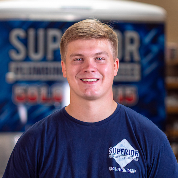 The Superior Plumbing & Rooter LLC intern Hunter. He is wearing a navy company t-shirt. Hunter is blond and standing in front of a blurry company wrapped trailer.