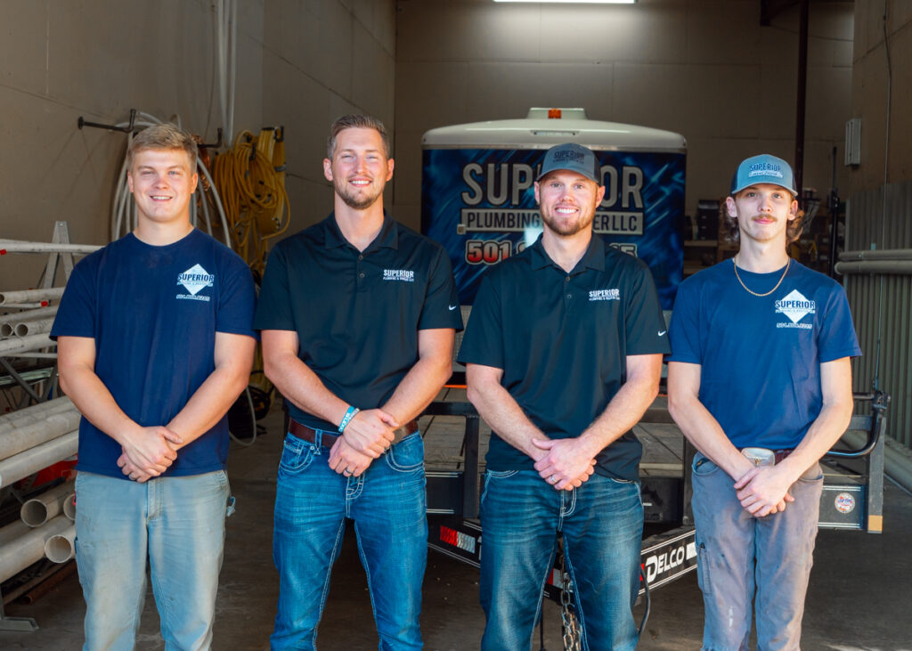 Two full time Superior Plumbing & Rooter LLC employees in black polos and two interns in blue company t-shirts. They pose clasping their hands in front of them in the garage in front of the company trailer.