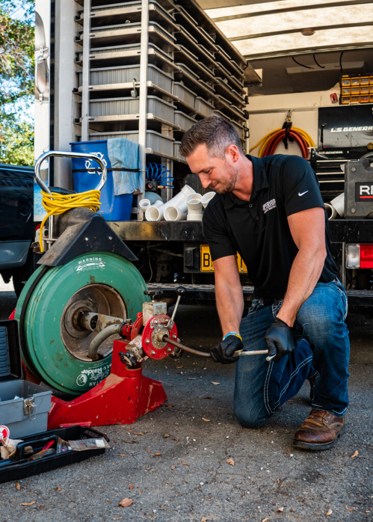 A Superior Plumbing & Rooter employee kneeling on the pavement and putting pressure on a rusty metal pipe protruding from a small red machine.