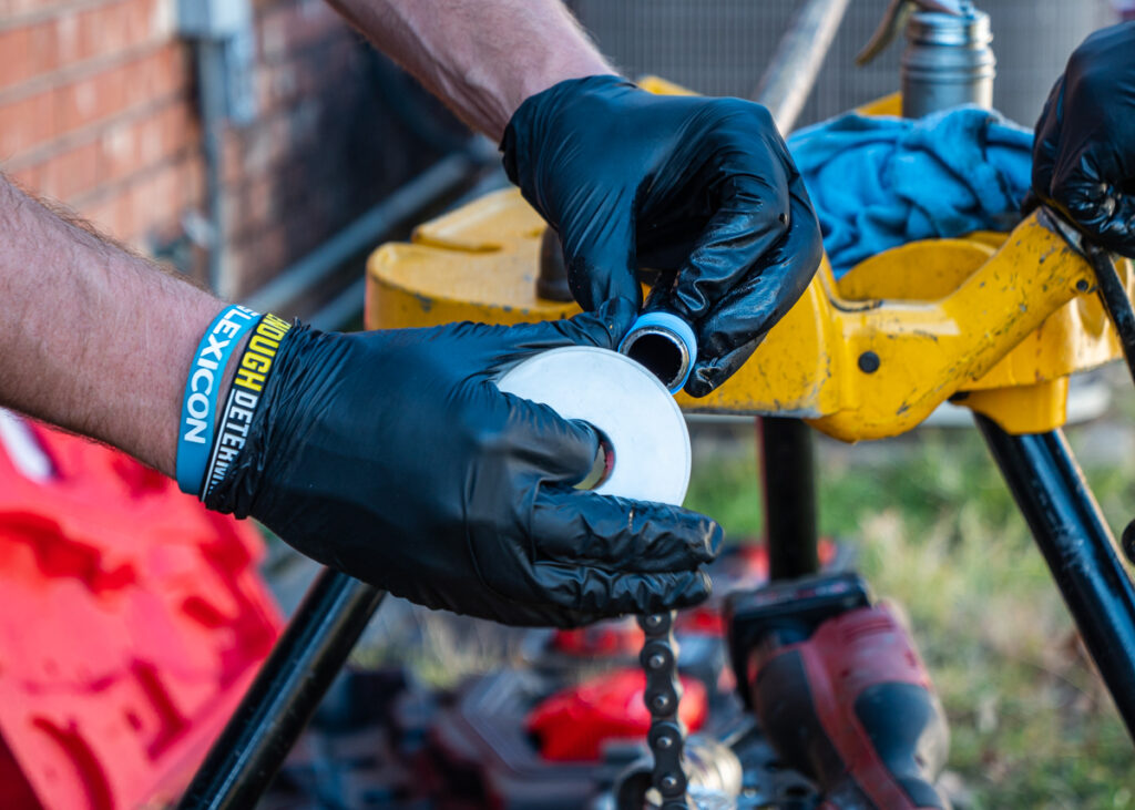 Two black-gloved hands hold the end of a pipe and a thick white circle with a hole in the middle. The pipe is resting on a yellow metal tool table.